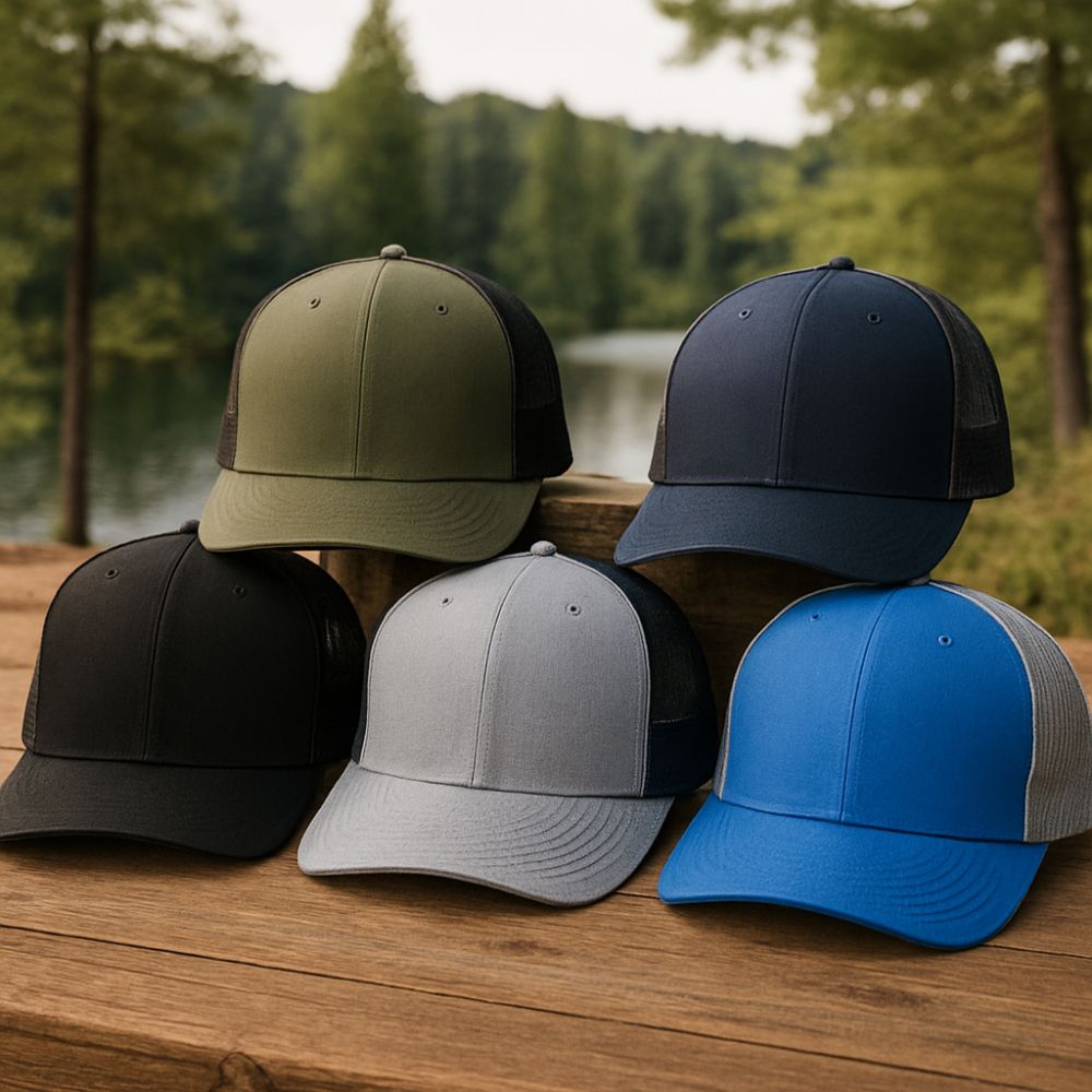 Assortment of five Richardson-style trucker hats in olive green, navy, black, grey, and royal blue, arranged on a wooden bench beside a scenic forest lake.